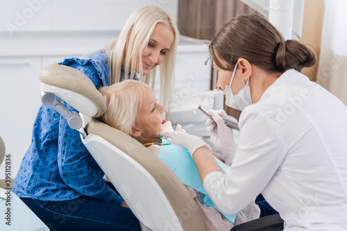 A child with a mother at a dentist s. The girl lies in the chair  with her mother behind. Procedure of dental hygiene.