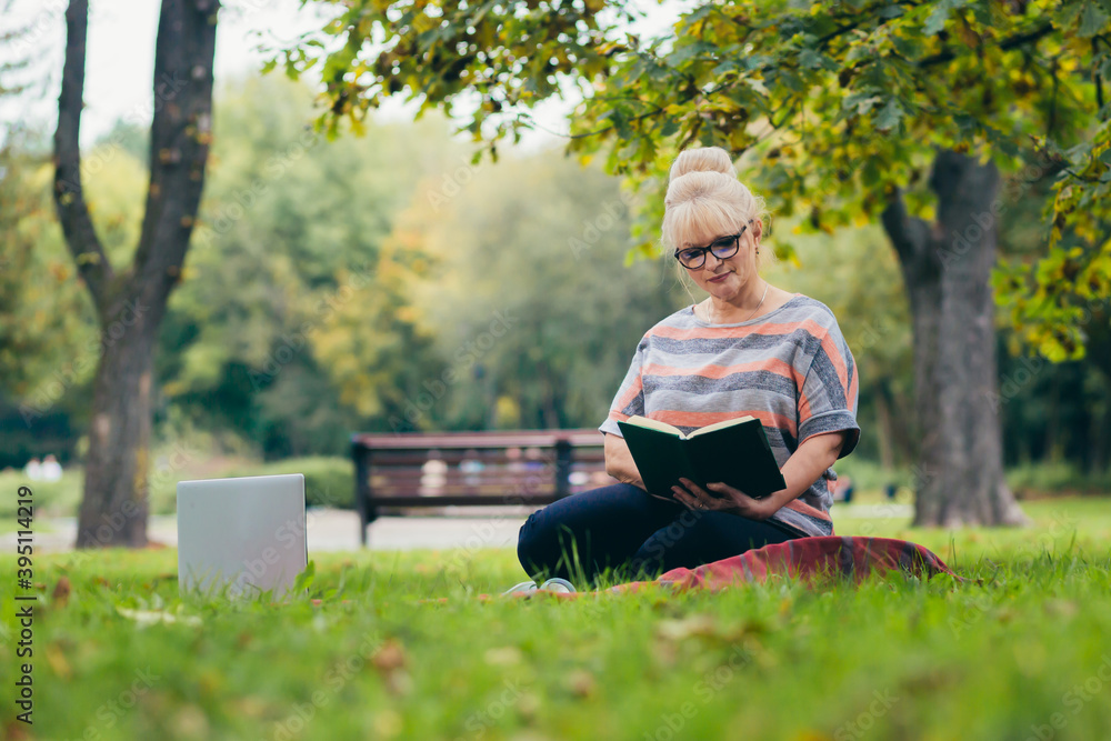 Senior woman resting in the park sitting on the grass, using phone and laptop, and reading a book Senior woman resting in the park sitting on the grass, using phone and laptop, and reading a book