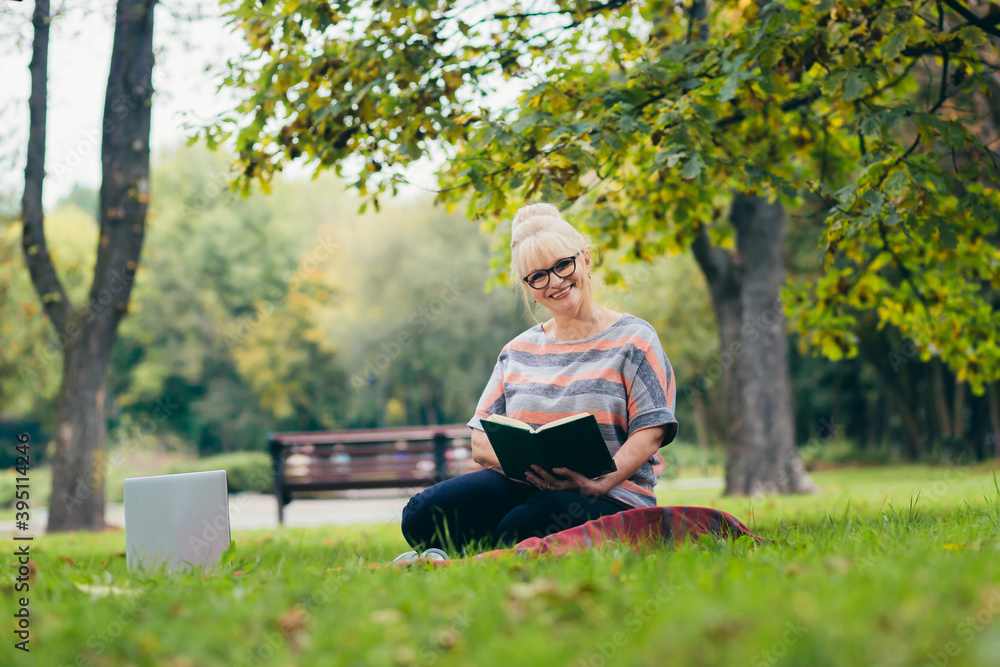 Senior woman resting in the park sitting on the grass, using phone and laptop, and reading a book Senior woman resting in the park sitting on the grass, using phone and laptop, and reading a book