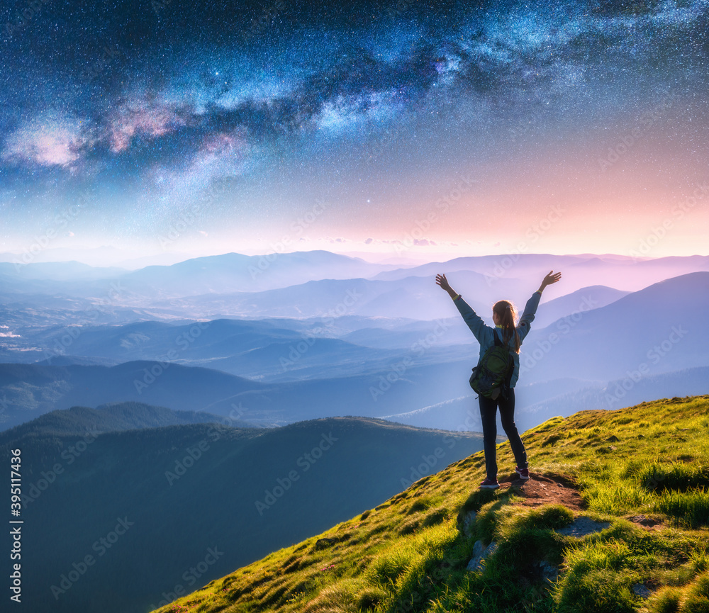 Happy young woman on the mountain peak and arched Milky Way over mountains at night. Landscape with girl, blue starry sky, Milky Way Arch, green grass, hills in fog. Space and galaxy. Sky with stars
