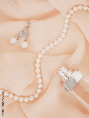 Women wedding jewelry. Necklace and earrings of natural pearls and unfocused bottle of perfume on gentle chiffon fabric as background. Luxury wedding background. Soft and delicate wavy pastel material