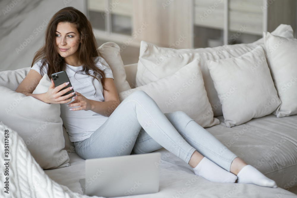 Brunette in a beautiful bright apartment. portrait of a woman sitting on the couch and taking online courses in a foreign language on the phone gadget device through the social network app. Brunette in a beautiful bright apartment. portrait of a woman sitting on the couch and taking online courses in a foreign language on the phone gadget device through the social network app.