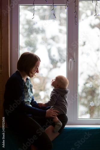 Young woman and her small child sitting on the window sill. Window is decorated for New Year. Snowy forest is outside. Christmas eve and family time concept