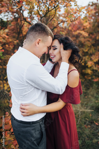A stylish respectable guy in a white shirt and trousers and a beautiful girl in an expensive long dress stand and laugh, hugging against the background of red, yellow bushes with leaves.