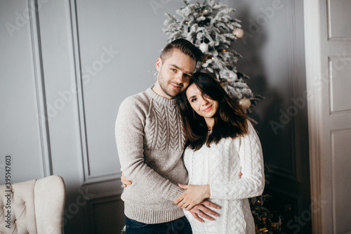 Adorable marriage couple stand near Christmas tree at home, caring husband hug pregnant wife, young family excited about parenthood, enjoy happiness, winter holidays concept