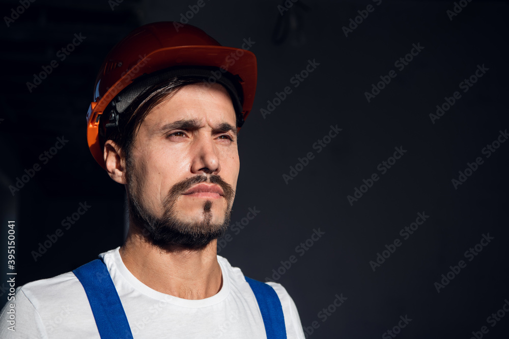 The worker puts an orange hard hat on his head. He is dressed in overalls and gloves. Close up The worker puts an orange hard hat on his head. He is dressed in overalls and gloves. Close up