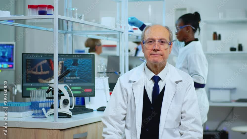 Senior chemist sitting in modern equipped lab looking satisfied at camera. Multiethnic team examining virus evolution using high tech and chemistry tools for scientific research, vaccine development Senior chemist sitting in modern equipped lab looking satisfied at camera. Multiethnic team examining virus evolution using high tech and chemistry tools for scientific research, vaccine development