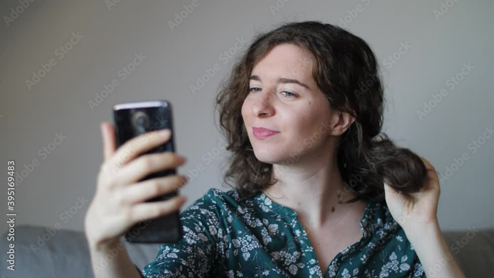 A beautiful happy young woman taking selfies in living room with phone. Attractive millennial girl with exotic shirt taking pictures of herself during quarantine at home in front of a white wall. A beautiful happy young woman taking selfies in living room with phone. Attractive millennial girl with exotic shirt taking pictures of herself during quarantine at home in front of a white wall.