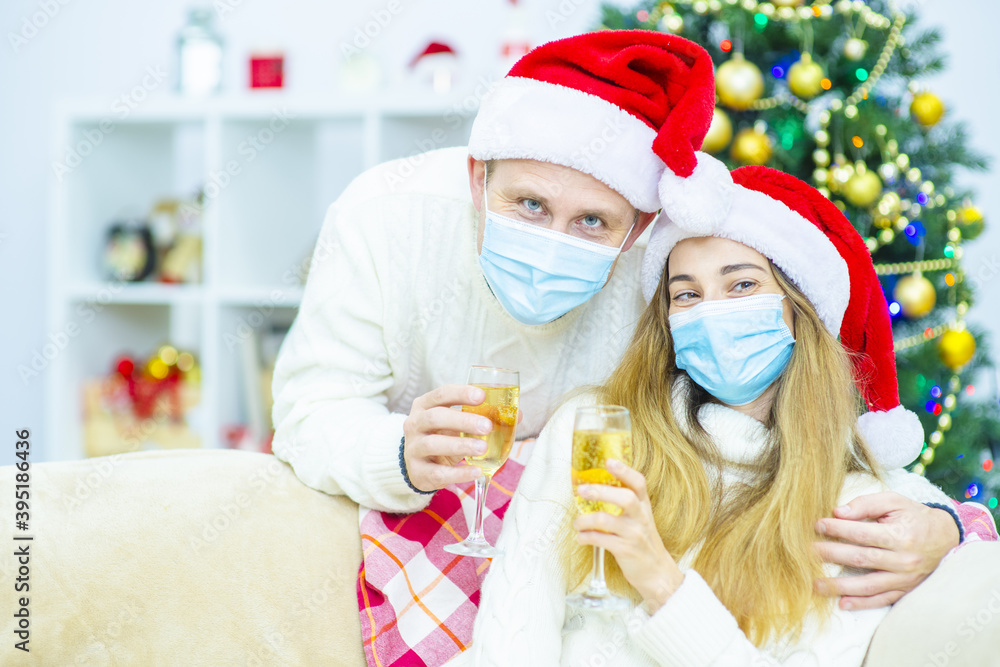 Fototapeta premium Young married couple with medical masks wearing santa hats with champagne in their hands wish each other a Merry Christmas (New Year)