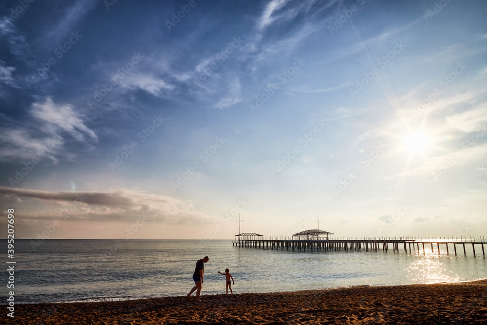 Fototapeta premium View from sand beach to water of sea, waves, pier and black silhouette of person in a nice day or evening with blues sky, bright sun and white clouds.