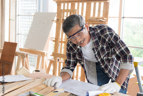 Senior asian old man carpenter wearing glasses and gloves protection check blueprint plan for building diy furniture in workshop