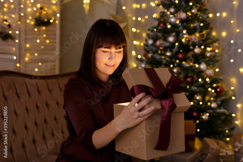 Christmas and magic concept - portrait of young woman opening magical gift box in decorated room with Christmas tree and led lights