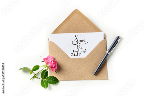 Save the date lettering on an invitation in a brown kraft envelope, shot from above on a white background with a pink rose