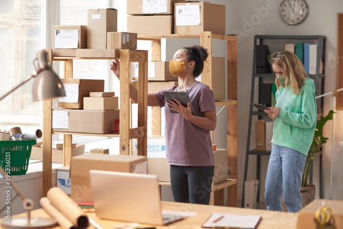 Two women in masks using digital tablet to check parcels on the shelves before delivery