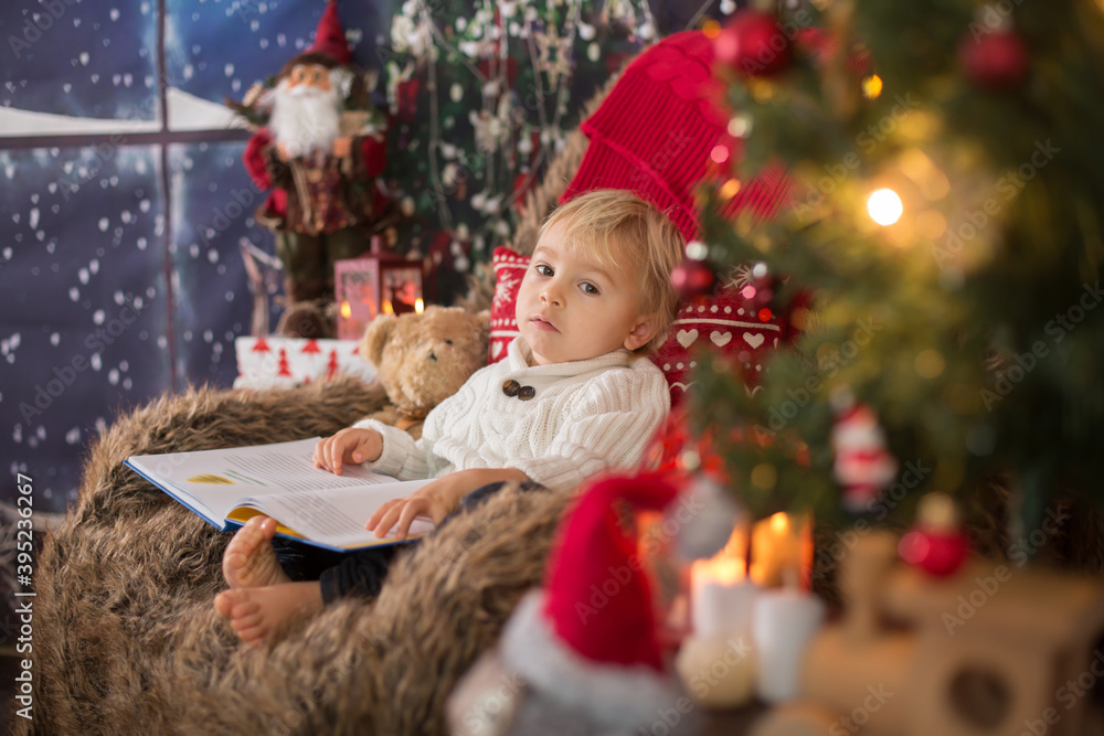 Cute toddler boy, sitting around christmas tree in cozy chair, reading book Cute toddler boy, sitting around christmas tree in cozy chair, reading book