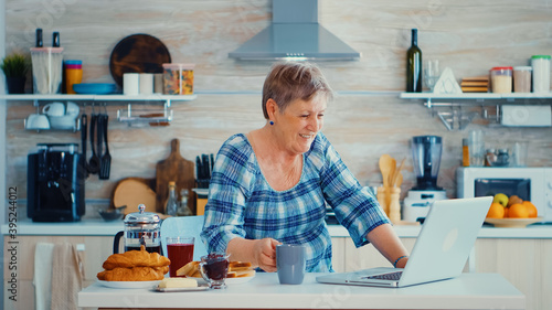 Senior woman drinking coffee and waving during a video conference with family in kitchen while having breakfast. Elderly person using internet online chat technology video webcam making a video call