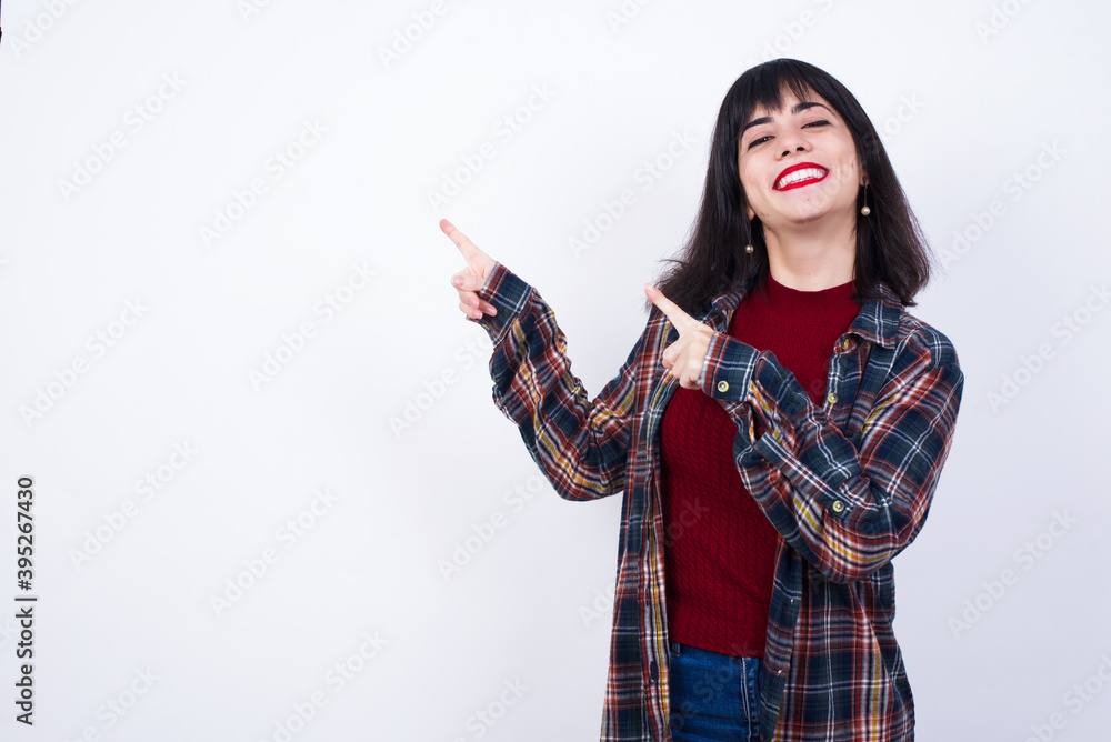 Positive Young beautiful Caucasian woman wearing plaid shirt against white wall, with beaming smile pointing with two fingers and looking on empty copy space. Advertisement concept. Positive Young beautiful Caucasian woman wearing plaid shirt against white wall, with beaming smile pointing with two fingers and looking on empty copy space. Advertisement concept.