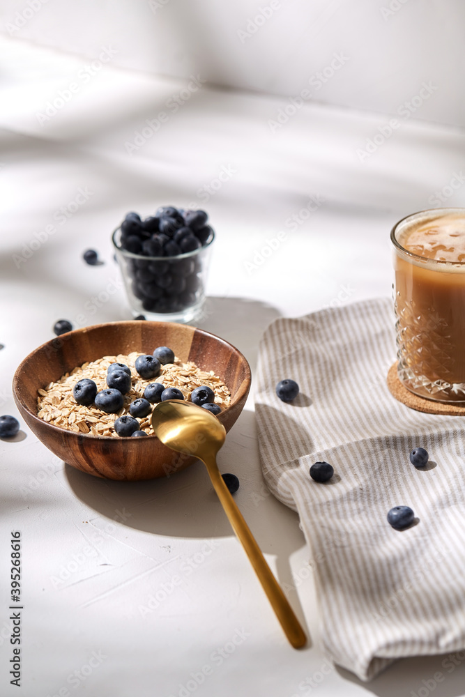 food, drinks and breakfast concept - oatmeal in wooden bowl with blueberries and spoon, glass of ice coffee on cork drink coaster and kitchen towel food, drinks and breakfast concept - oatmeal in wooden bowl with blueberries and spoon, glass of ice coffee on cork drink coaster and kitchen towel