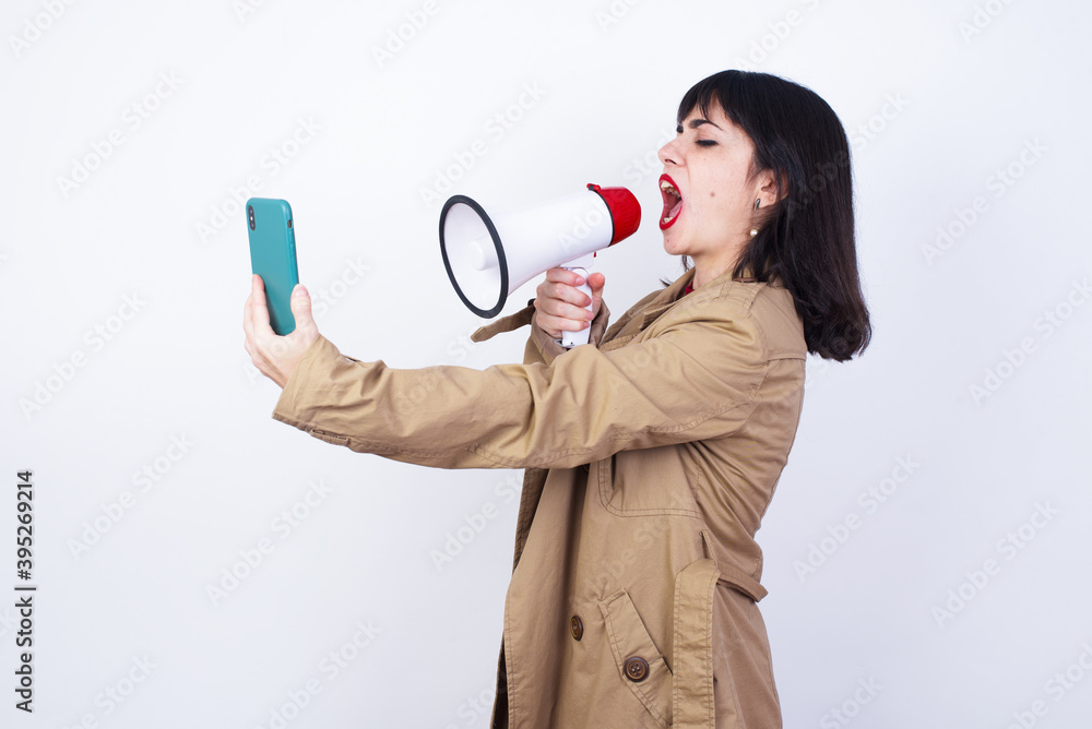 Young beautiful caucasian businesswoman standing against white background, communicates shouting at smartphone with megaphone. Marketing and sales concept.