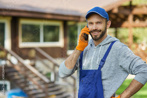 Happy young male builder in uniform smiling at camera while talking on the phone, standing outdoors at cottage construction site