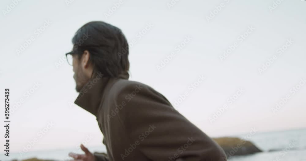 Young man in brown coat and glasses runs on near beach looking back. Run away from someone. Young man in brown coat and glasses runs on near beach looking back. Run away from someone.