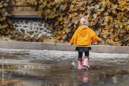 A joyful little girl in a yellow raincoat and pink rubber boots jumps on puddles with splashes and rejoices. Park, nature, outdoors. Universal Children's Day.