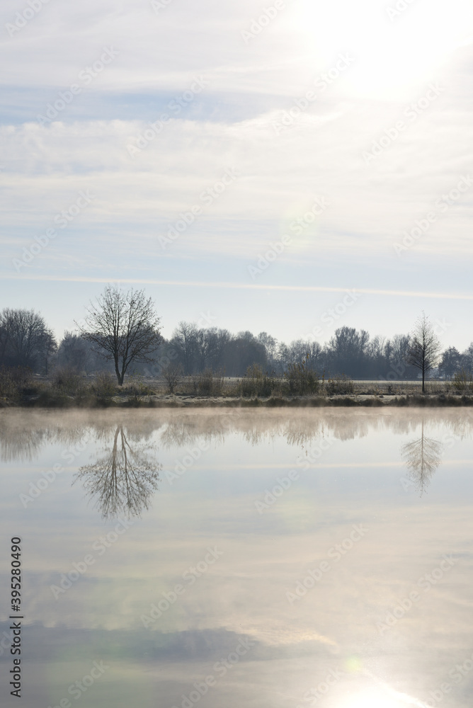 Scenic shot of a lake in winter, above which the haze is slowly rising, with trees in the background