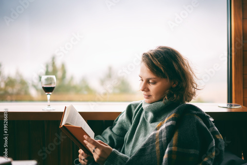 Fotografie Young woman in green sweater reads a book and drinking wine near window in count