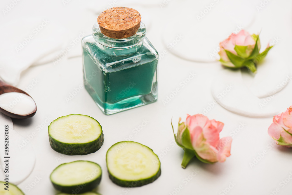 cucumber slices near corked bottle of homemade lotion, tea roses, and cotton pads on white blurred 