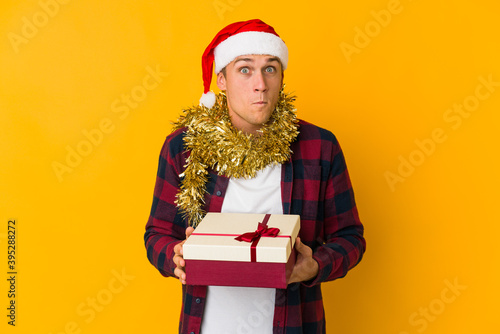 Young caucasian man with christmas hat holding a present isolated on yellow background