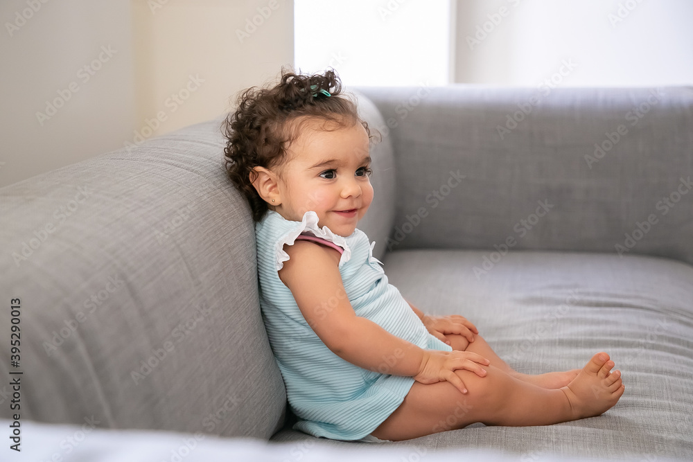 Joyful dark curly haired baby girl wearing pale blue cloth, sitting on grey couch at home, looking away and smiling. Kid at home and childhood concept Joyful dark curly haired baby girl wearing pale blue cloth, sitting on grey couch at home, looking away and smiling. Kid at home and childhood concept