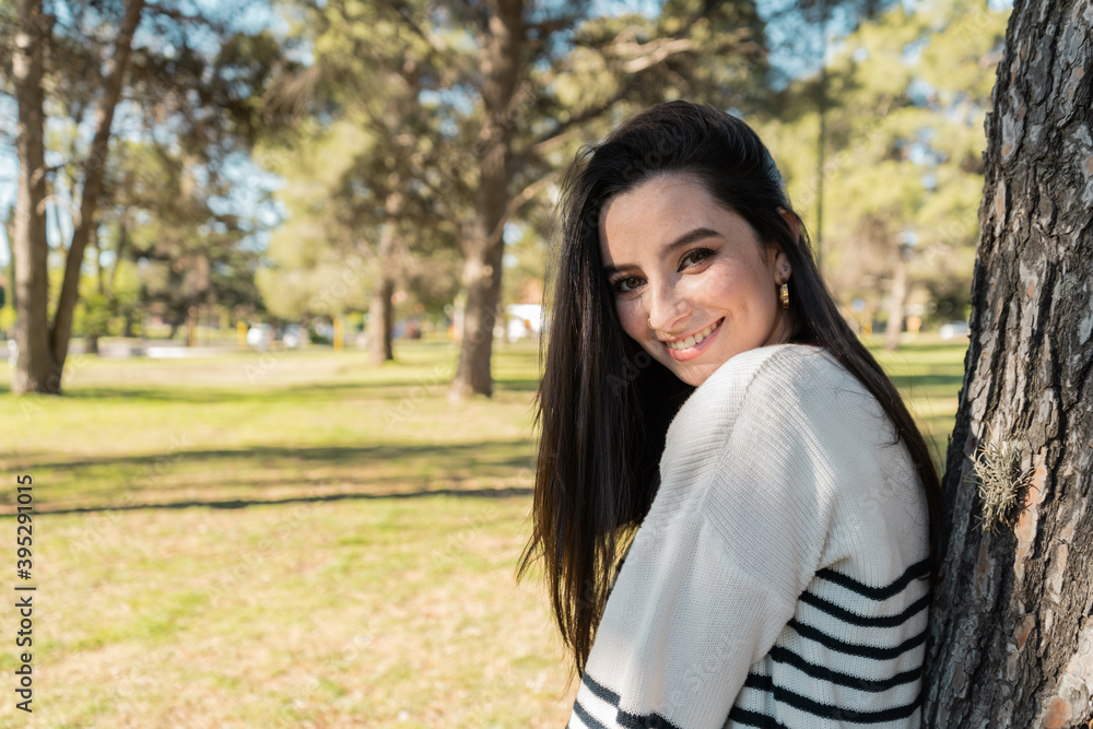 Beautiful black-haired woman in a park, leaning on a tree and looking at the camera Beautiful black-haired woman in a park, leaning on a tree and looking at the camera