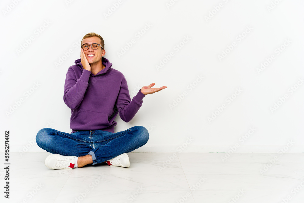 Young caucasian man sitting on the floor holds copy space on a palm, keep hand over cheek. Amazed and delighted.