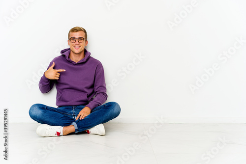 Young caucasian man sitting on the floor person pointing by hand to a shirt copy space, proud and confident © Asier