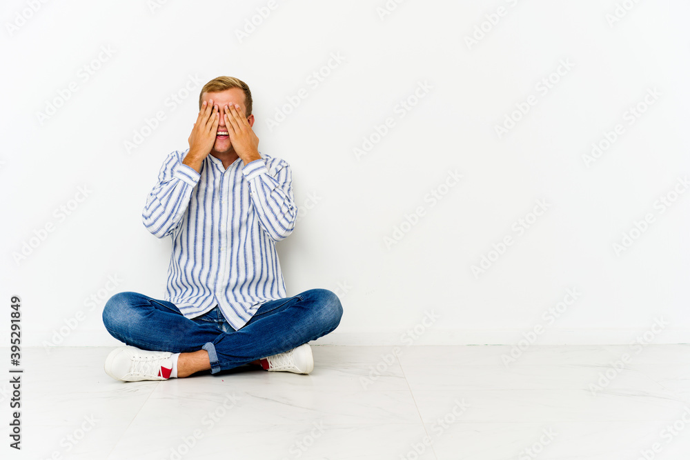 Young caucasian man sitting on the floor covers eyes with hands, smiles broadly waiting for a surprise. Young caucasian man sitting on the floor covers eyes with hands, smiles broadly waiting for a surprise.