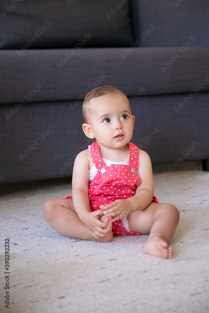 Lovely little baby sitting on carpet barefoot in living room. Cute thoughtful girl in red dungarees shorts looking at someone and touching foot. Weekend, childhood and being at home concept Lovely little baby sitting on carpet barefoot in living room. Cute thoughtful girl in red dungarees shorts looking at someone and touching foot. Weekend, childhood and being at home concept