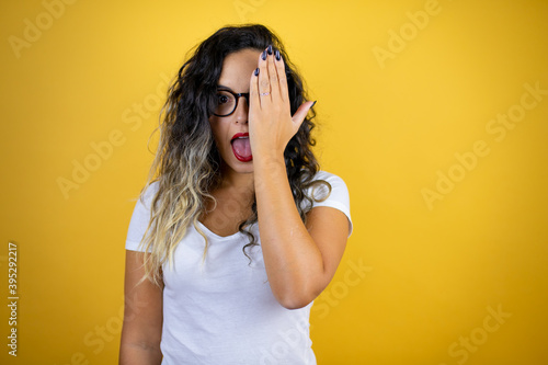 Young beautiful woman wearing casual white t-shirt over isolated yellow background covering one eye with hand, confident smile on face and surprise emotion.