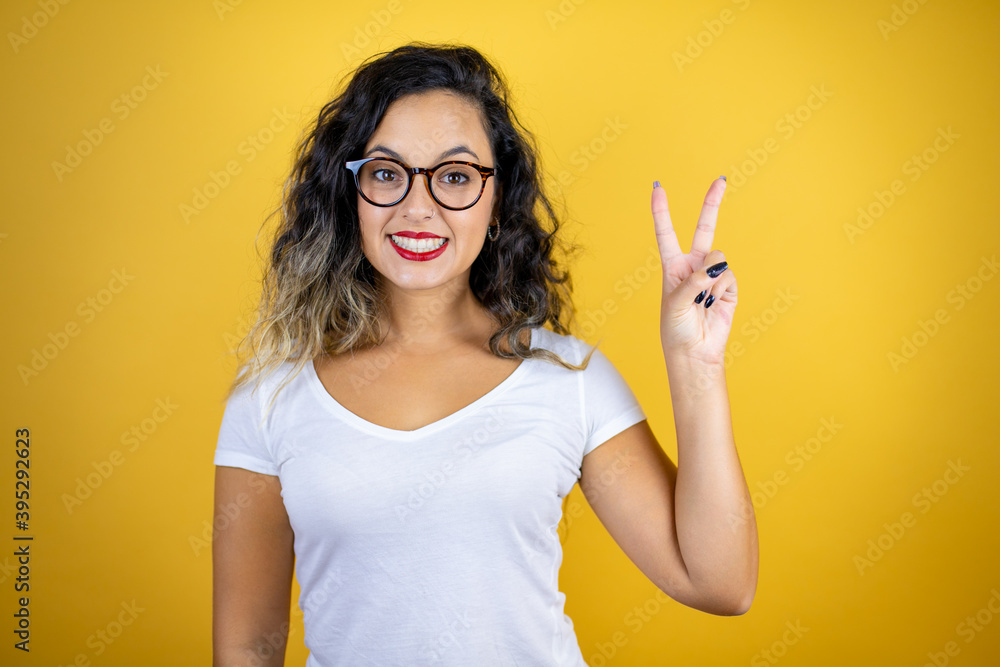 Young beautiful woman wearing casual white t-shirt over isolated yellow background showing and pointing up with fingers number two while smiling confident and happy