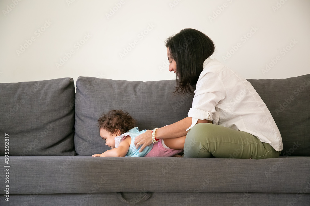 Happy black haired mom cuddling cute baby daughter on grey couch. Side view. Parenthood and childhood concept