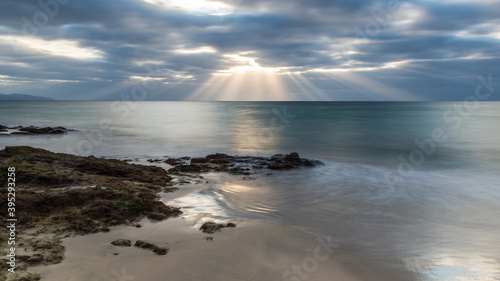 Dramatic sunrise by the sea. A seascape at sunrise with sun beams passing through coloured clouds. At foreground the sea shore with reflections of the sky in the puddles among the rocks.