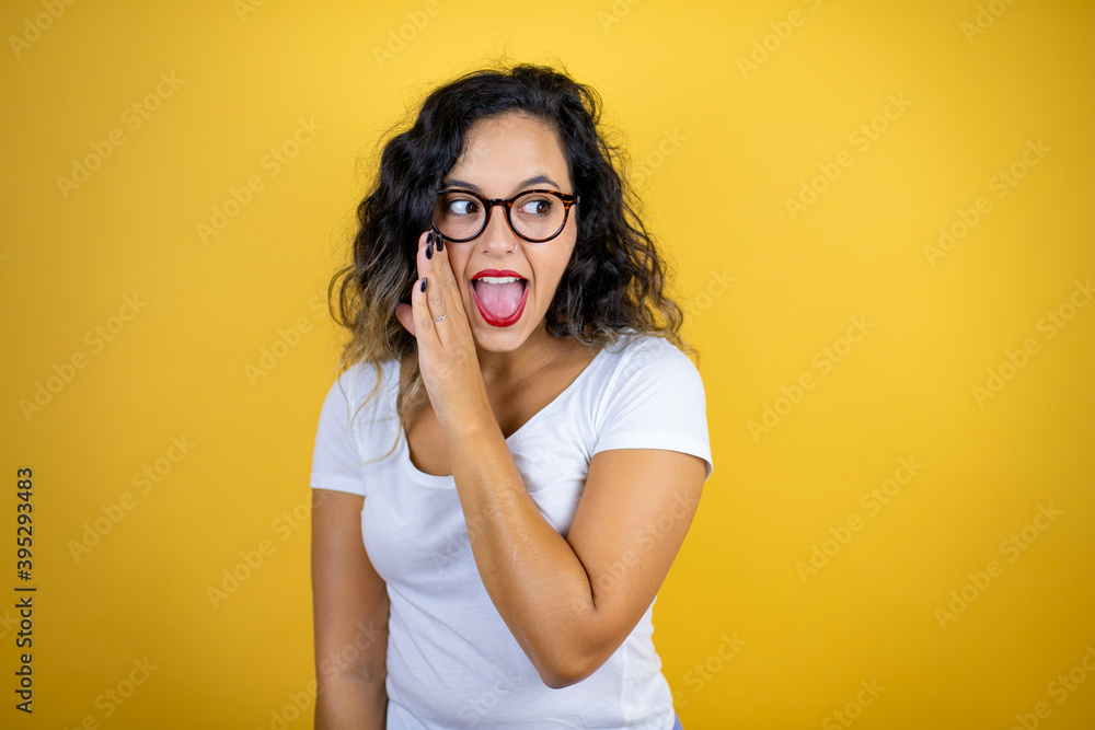 Young beautiful woman wearing casual white t-shirt over isolated yellow background with her hand on mouth telling secret rumor, whispering malicious talk conversation Young beautiful woman wearing casual white t-shirt over isolated yellow background with her hand on mouth telling secret rumor, whispering malicious talk conversation