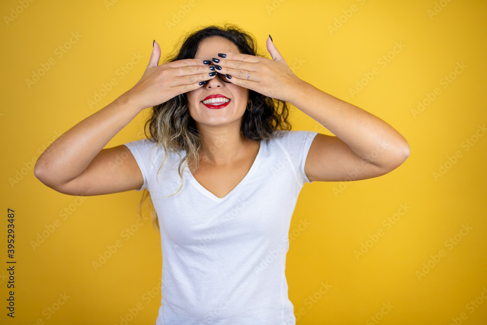 Fototapeta premium Young beautiful woman wearing casual white t-shirt over isolated yellow background covering eyes with hands smiling cheerful and funny. Blind concept.