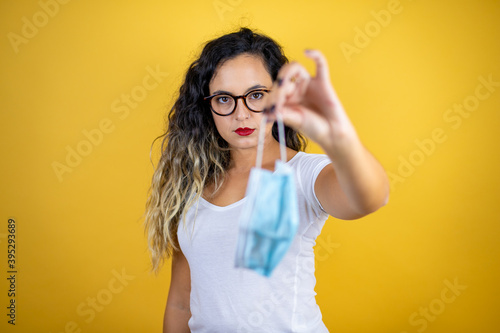 Young beautiful woman wearing casual white t-shirt over isolated yellow background serious showing the mask