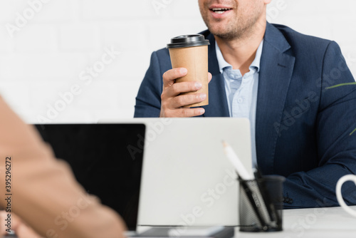 Cropped of office worker with paper cup talking, while sitting at table with laptops on blurred foreground