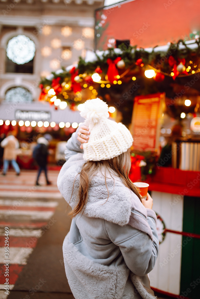 Young woman relaxing drinking coffee in  Festive Christmas fair. Smiling woman in winter style clothes walking in Christmas market. Winter holidays. Lights around.