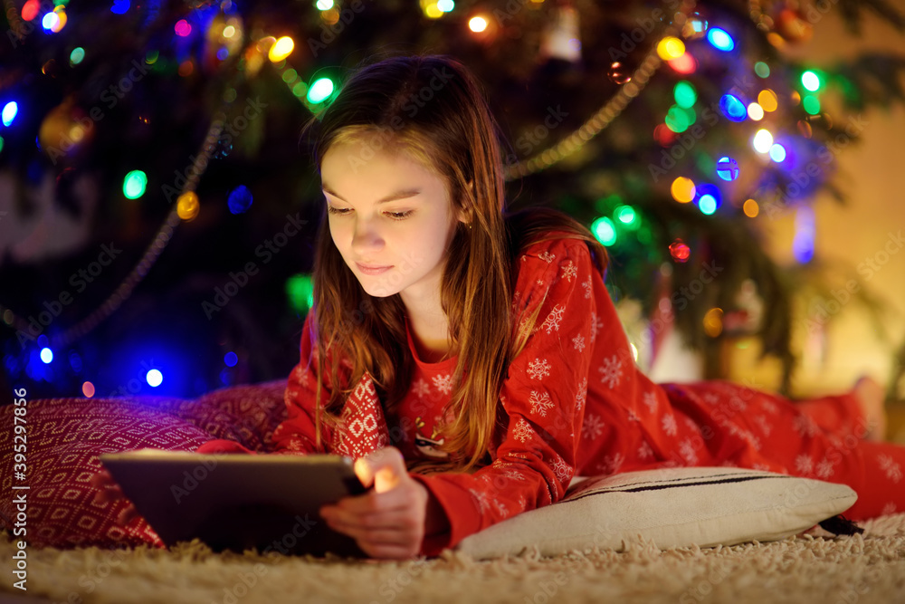 Adorable young girl using a tablet pc at home by a fireplace in warm and cozy living room on Christmas eve. Winter evening with family and kids. Adorable young girl using a tablet pc at home by a fireplace in warm and cozy living room on Christmas eve. Winter evening with family and kids.