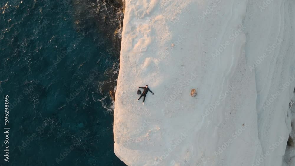 Aerial top down shot rising over a young man lying on a huge volcanic rock in Sarakiniko beach, Greece Aerial top down shot rising over a young man lying on a huge volcanic rock in Sarakiniko beach, Greece