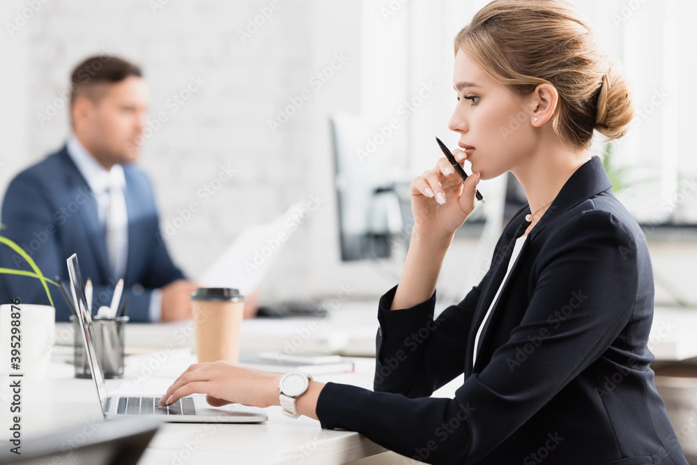 Side view of businesswoman with pen typing on laptop, while sitting at workplace on blurred background Side view of businesswoman with pen typing on laptop, while sitting at workplace on blurred background