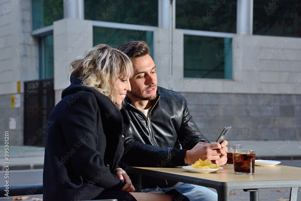 Two young friends consult the mobile sitting on the terrace of a bar