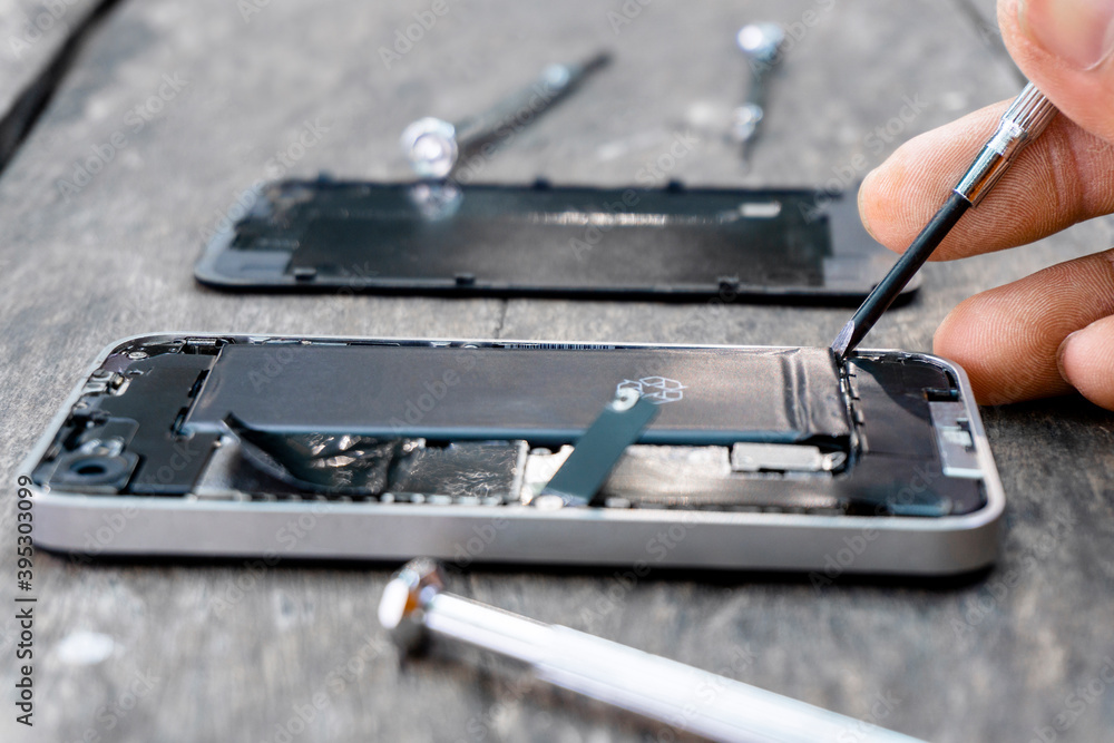 The technician holding a screwdriver a mobile phone repair Closeup inside cell phone with a fixing battery from a broken service shop center on wood table. smartphone repairs maintenance concept. The technician holding a screwdriver a mobile phone repair Closeup inside cell phone with a fixing battery from a broken service shop center on wood table. smartphone repairs maintenance concept.